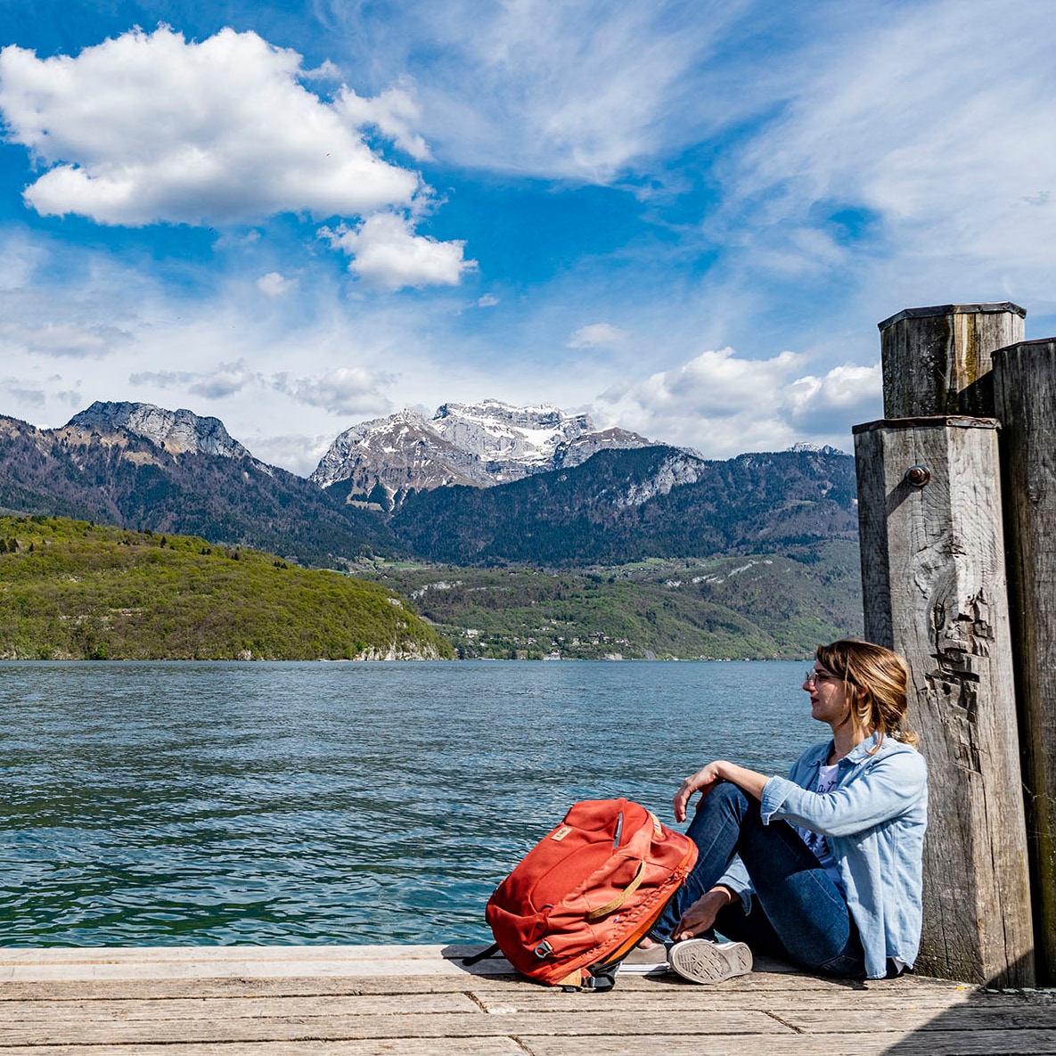 Une femme est assise sur le ponton en bois au bord du lac Annecy avec son sac à dos NEO à ses pieds